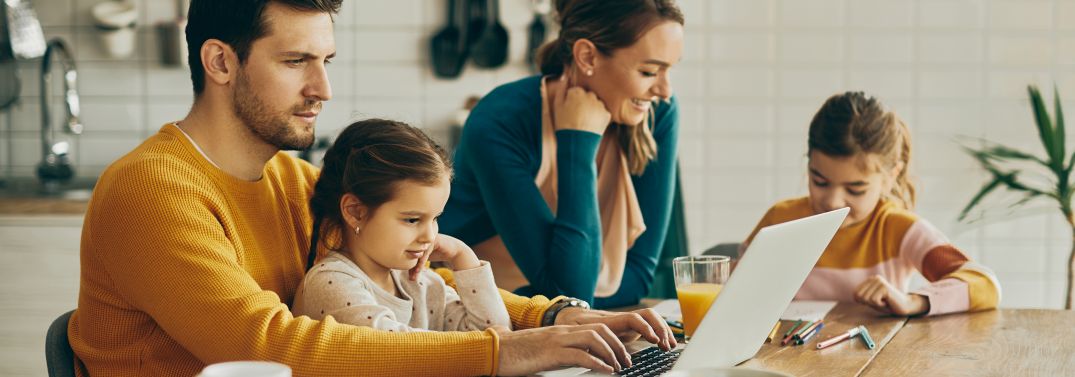 Junge Familie sitzt zusammen am Tisch mit Hausaufgaben und PC